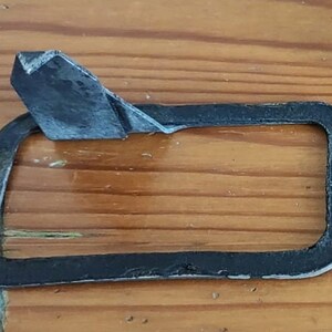 May include: A black gasket with a rectangular centre and rounded corners. The gasket has two holes and a folded-over section at the top. The object is on a wooden surface.