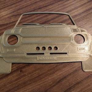 May include: A gold-colored, 3D-printed car front with round headlights, a grille with three holes, and the name "CARVEN" on the license plate. The car's windshield and front bumper are also visible.
