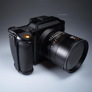 Op de afbeelding: Een zwarte Hasselblad camera met een 55mm lens. De camerabody heeft een getextureerde grip en een oranje knop. De lens heeft witte en oranje markeringen. Het woord "Hasselblad" staat op de bovenkant van de camera.