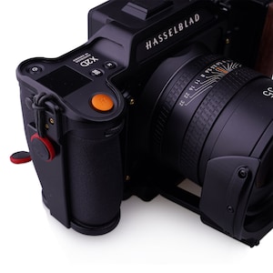 K&ouml;nnte beinhalten: Eine schwarze Hasselblad X2D Kamera mit einem orangen Knopf und einem schwarzen Objektiv mit einer Tiefensch&auml;rfenskala. Die Kamera hat einen schwarzen Lederriemen, der an der Seite befestigt ist.