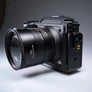 May include: A black Hasselblad camera with a large lens. The camera body and lens are black, with the Hasselblad logo visible. The lens has measurement markings. The camera is mounted on a black base.