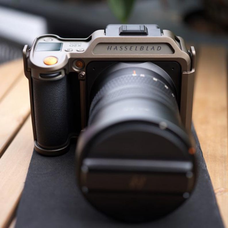 Może przedstawiać: Aparat Hasselblad średniego formatu z dużym obiektywem. Korpus aparatu jest jasnoszary z napisem "Hasselblad" na g&oacute;rze. Obiektyw jest czarny, a aparat ma czarny uchwyt.