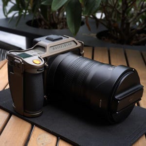 May include: A professional digital camera with a large black lens and the brand name "Hasselblad" printed on the top. The camera body is a muted bronze color with a black grip and a small orange button. The lens is a matte black.