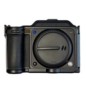 May include: A black Hasselblad X System camera with a prominent lens and grip. The camera body is dark gray with the brand name across the top. A blue light is visible on the front.