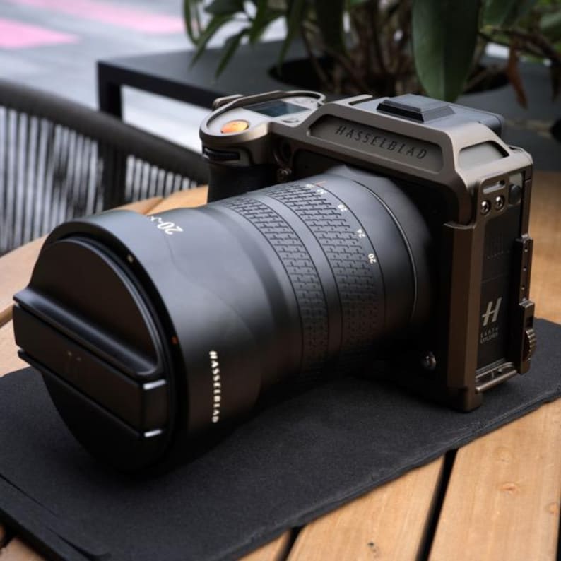 Może przedstawiać: Profesjonalny aparat Hasselblad z dużym czarnym obiektywem. Korpus aparatu jest w kolorze ciemnego brązu, z wytłoczonym logo marki na g&oacute;rze. Obiektyw ma oznaczenia pomiarowe. Aparat stoi na czarnej powierzchni.