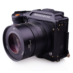 K&ouml;nnte beinhalten: Eine schwarze Hasselblad-Kamera mit einem gro&szlig;en Objektiv. Das Objektiv hat einen schwarzen Lauf mit wei&szlig;em Text, der "HASSELBLAD" und "50mm" lautet. Der Kamerak&ouml;rper hat ein schwarzes und goldenes Design mit dem Wort "HASSELBLAD" in wei&szlig; auf der Oberseite.