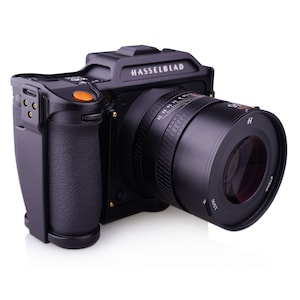 May include: A black Hasselblad camera with a black lens. The camera has a black grip and a small orange button on the top.