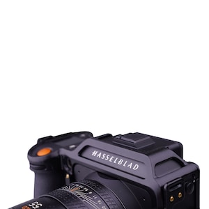 K&ouml;nnte beinhalten: Eine schwarze Hasselblad-Kamera mit einem gro&szlig;en Objektiv und einem orangefarbenen Knopf an der Seite. Die Kamera hat ein schwarzes Lederband und ein silbernes Typenschild mit der Aufschrift "HASSELBLAD".