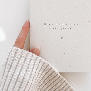 Könnte beinhalten: Ein weißes Notizbuch mit dem Text "Mutterpass" und "marie louise" darauf. Ein kleines Herz ist unter dem Text gezeichnet.