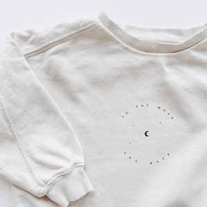 Könnte beinhalten: Ein cremefarbener Pullover mit einem Motiv eines Halbmondes und dem Text "To the moon and back" in einem Kreis.