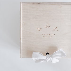 Könnte beinhalten: Eine helle Holzkiste mit einem weißen Band darum gebunden. Die Kiste hat ein schlichtes Design mit drei Vögeln und dem Text "JOHANNA 23.01.24" auf der Vorderseite.