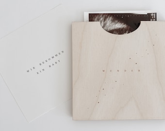 Puede incluir: Una caja de madera con un recorte para una foto de ecografía. La caja es de color marrón claro y tiene el texto "WIR BEKOMMEN EIN BABY" en la parte delantera. El texto "WUNDER" también está en la parte delantera de la caja.