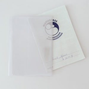 Könnte beinhalten: Eine transparente Plastikhülle für ein Dokument. Das Dokument ist ein weißes Blatt Papier mit einem blau-weißen Logo, auf dem "Mutterpass Gemeinsamer Bundesausschuss" steht. Das Dokument trägt eine handschriftliche Unterschrift und ein Datum.