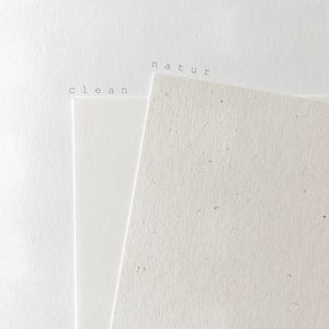 May include: A minimalist composition featuring three sheets of white paper. The paper on the right has a textured, speckled surface, while the other two are smooth. The words "clean" and "natur" are printed in a small, sans-serif font.
