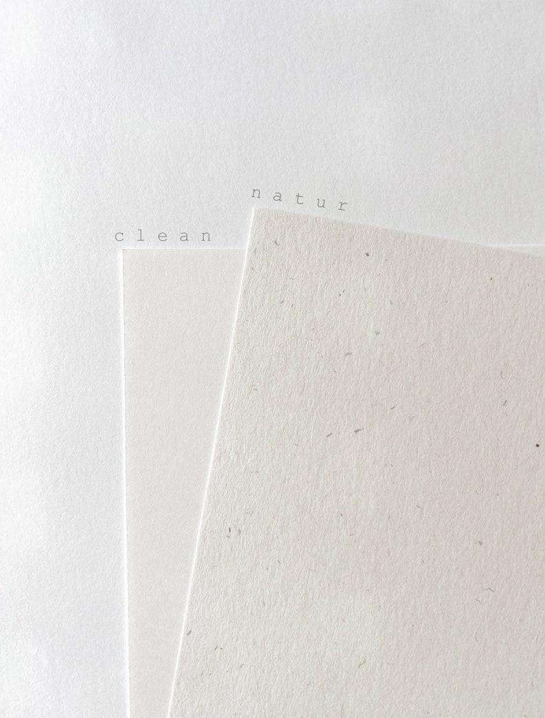 May include: A minimalist composition featuring three sheets of white paper. The paper on the right has a textured, speckled surface, while the other two are smooth. The words "clean" and "natur" are printed in a small, sans-serif font.