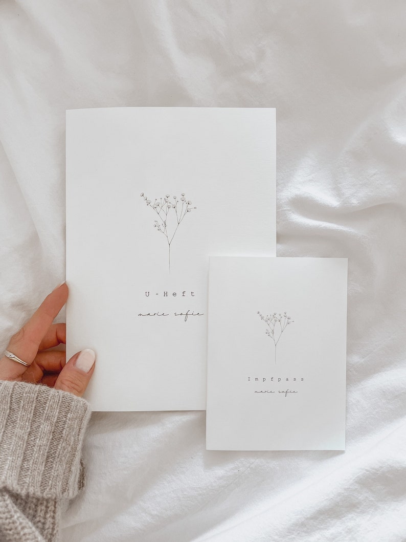 May include: Two white cards with minimalist floral illustrations and text. The larger card reads "U-Heft marie sofie", while the smaller one says "Impfpass marie sofie". The cards are on a white fabric background.