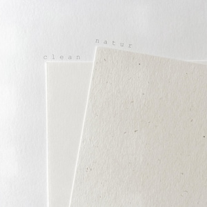 May include: Two sheets of paper are layered. The left sheet is smooth, bright white, and labeled with the word clean. The right sheet is off-white, textured with visible flecks, and labeled with the word natur. Both labels are in black.