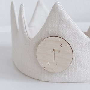 Könnte beinhalten: Eine weiße Stoffkrone mit einem daran befestigten Holzkreis. Der Kreis trägt die Zahl "1" und einen eingravierten Halbmond.