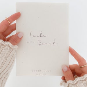 Könnte beinhalten: Eine weiße Karte mit dem Text "Liebe im Bauch" und "Sarah Jones 01.10.1992" in schwarzer Tinte.