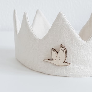 Könnte beinhalten: Eine weiße Leinenkrone mit einem ausgeschnittenen Holzschwan, der an der Vorderseite befestigt ist. Der Schwan trägt die Nummer "2".