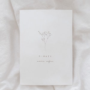 May include: A white card with a delicate floral illustration and the text "U-Heft marie sofie" in a minimalist style. The card is set against a soft, white fabric background.