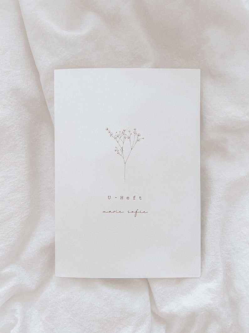 May include: A white card with a delicate floral illustration and the text "U-Heft marie sofie" in a minimalist style. The card is set against a soft, white fabric background.