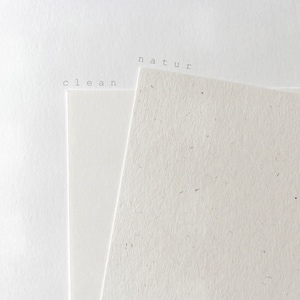 Puede incluir: Dos piezas de papel, una blanca y otra con una textura moteada. El papel blanco tiene la palabra "clean" impresa, y el papel moteado tiene la palabra "natur" impresa.