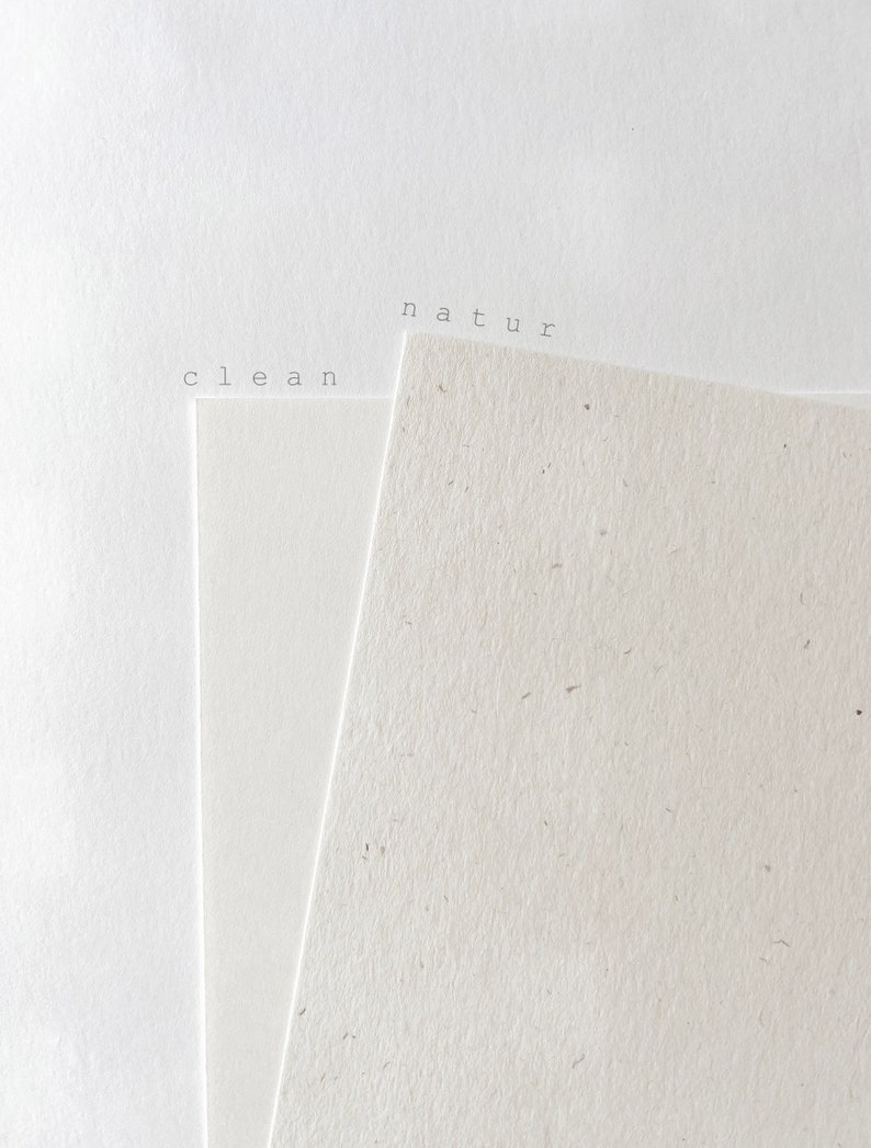 Puede incluir: Dos piezas de papel, una blanca y otra con una textura moteada. El papel blanco tiene la palabra "clean" impresa, y el papel moteado tiene la palabra "natur" impresa.