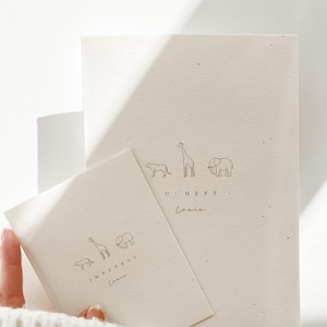 May include: Two off-white paper booklets with speckled texture. The larger booklet features line drawings of a cheetah, giraffe, and elephant above the text U HEFT and the name Louis. The smaller booklet features the same animals above IMPFPASS and Louis.