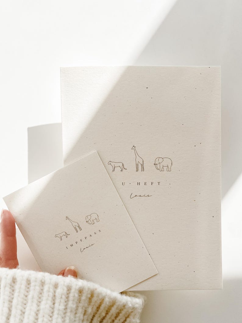May include: Two off-white paper booklets with speckled texture. The larger booklet features line drawings of a cheetah, giraffe, and elephant above the text U HEFT and the name Louis. The smaller booklet features the same animals above IMPFPASS and Louis.