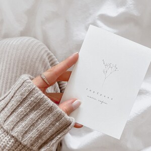May include: A white card with the word "Impfpass" and other text, along with a delicate floral illustration. The card is held against a background of white fabric and a beige sweater.