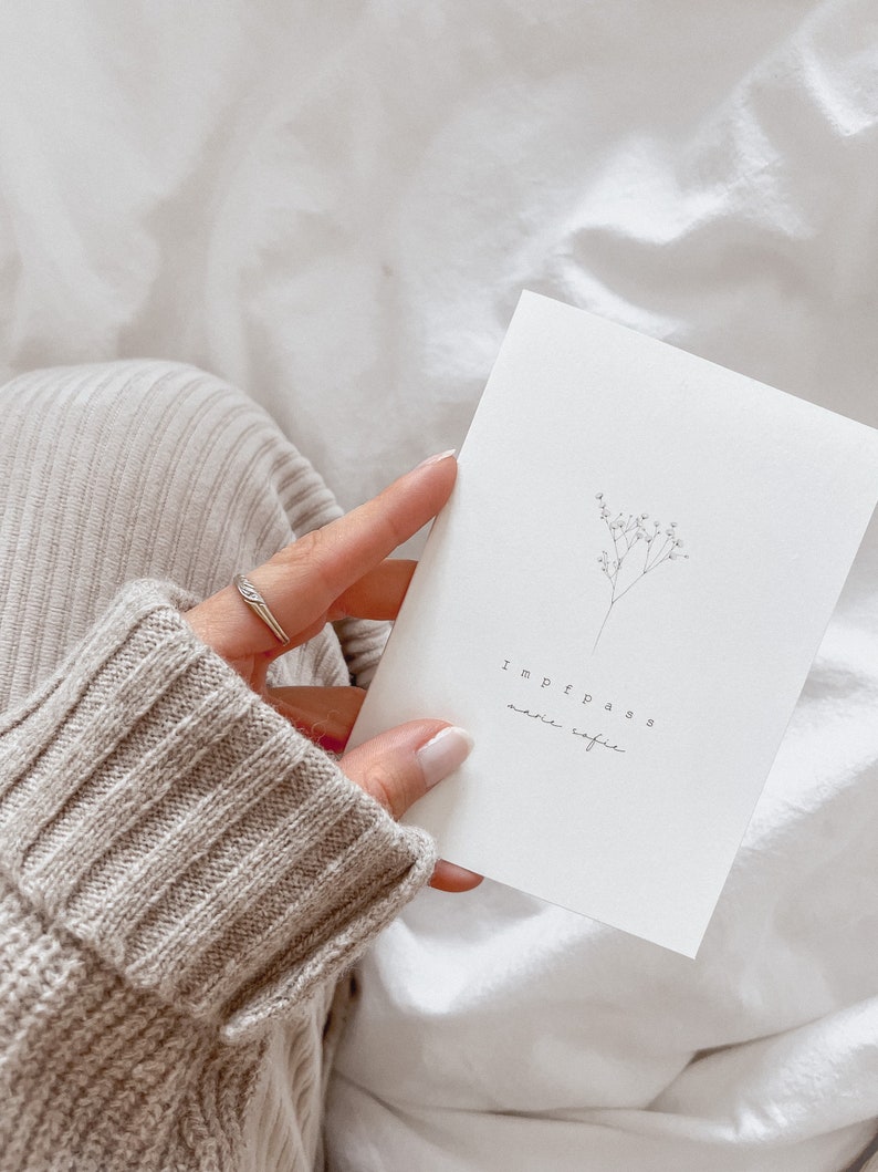 May include: A white card with the word "Impfpass" and other text, along with a delicate floral illustration. The card is held against a background of white fabric and a beige sweater.