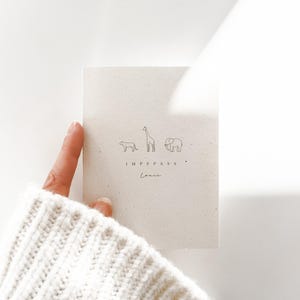 May include: A rectangular off-white card featuring a minimalist line drawing of a lion, a giraffe, and an elephant. Below the animals, the text reads IMPFPASS and the name Louis in a cursive script, printed in a dark, thin font.