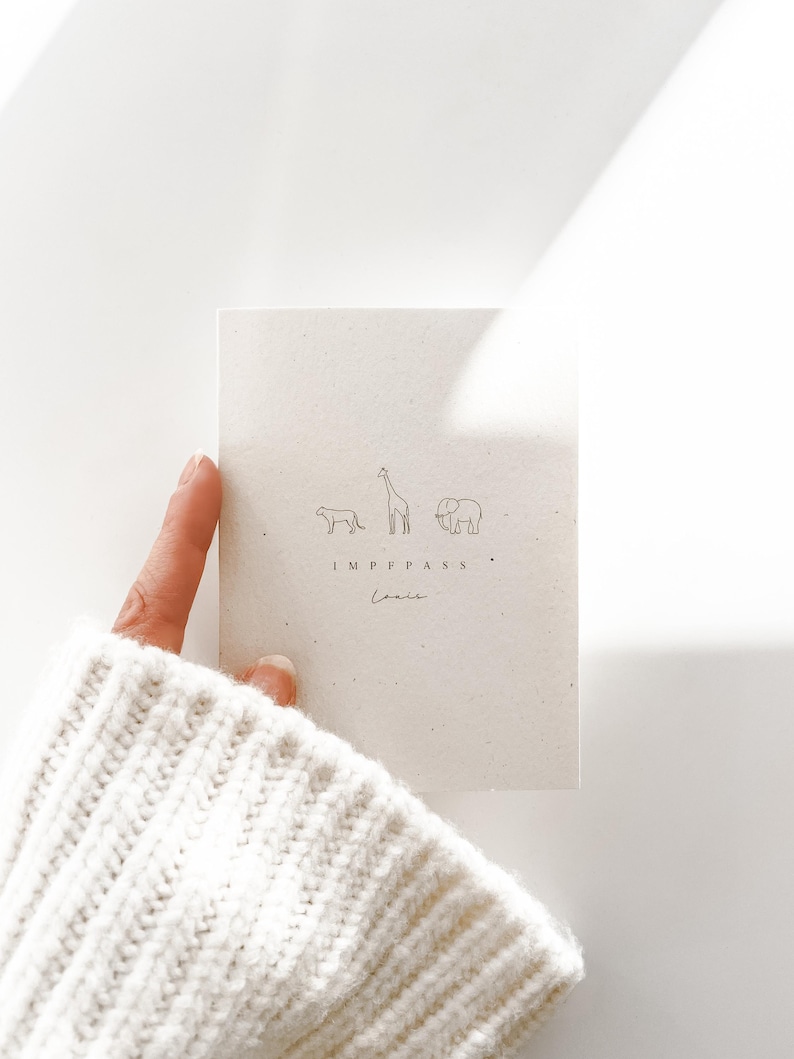 May include: A rectangular off-white card featuring a minimalist line drawing of a lion, a giraffe, and an elephant. Below the animals, the text reads IMPFPASS and the name Louis in a cursive script, printed in a dark, thin font.