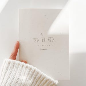 May include: A cream-colored booklet featuring a minimalist line drawing of a cheetah, a giraffe, and an elephant. Below the animals, the text reads U HEFT and the name Louie is written in a cursive script. The cover has a speckled texture.