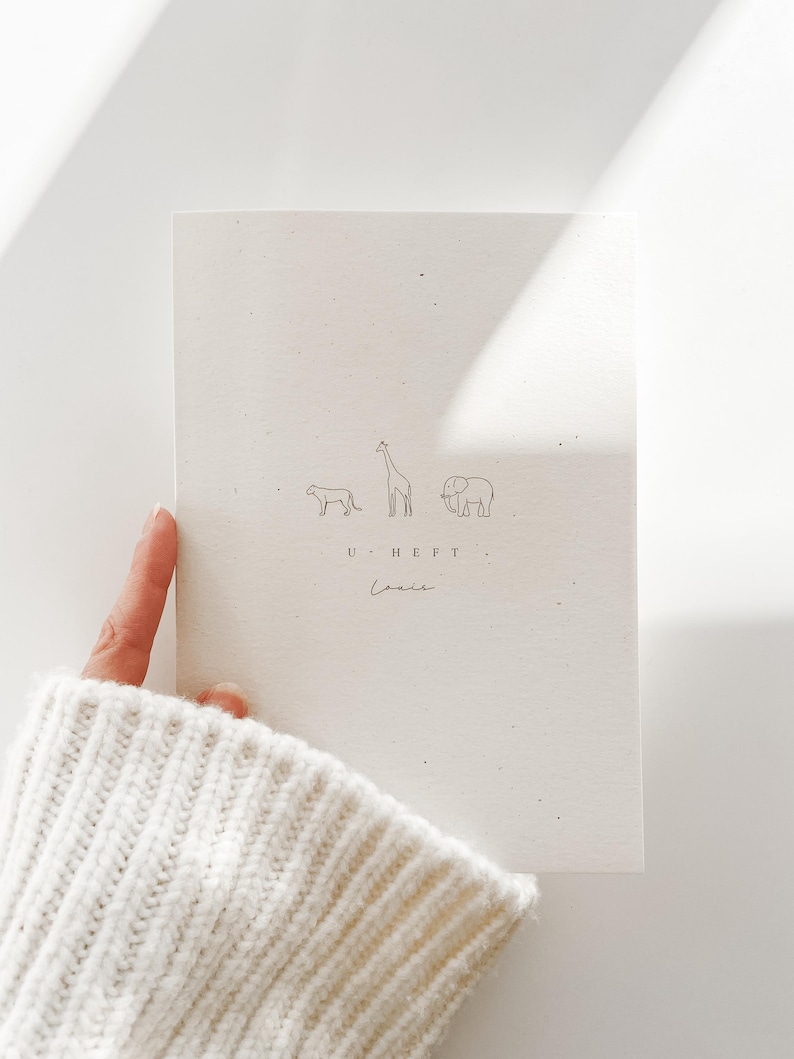 May include: A cream-colored booklet featuring a minimalist line drawing of a cheetah, a giraffe, and an elephant. Below the animals, the text reads U HEFT and the name Louie is written in a cursive script. The cover has a speckled texture.
