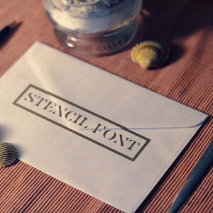 Puede incluir: Un sobre blanco con un borde negro alrededor del texto "STENCIL FONT". El sobre est&aacute; sobre una superficie marr&oacute;n texturizada con una concha y un bol&iacute;grafo.