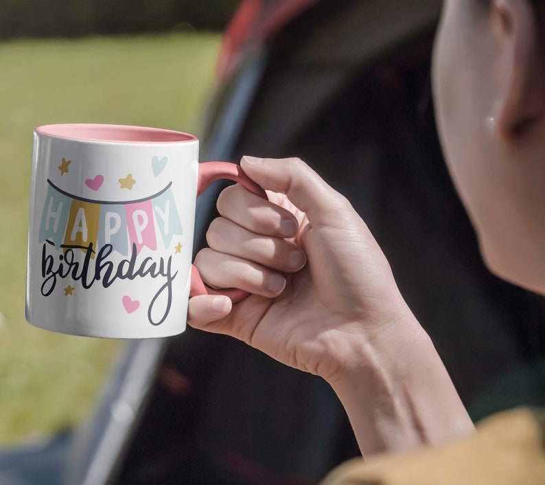 May include: White ceramic mug with a pink interior and handle. The mug features the words "Happy Birthday" in a script font, with a banner design in pastel colors and small star and heart accents. The mug is held by a person.