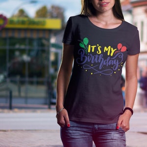 May include: Black t-shirt with the text "IT'S MY Birthday" in a colorful design featuring balloons. The text is in yellow, purple, and blue, with green and red balloons. The shirt is worn by a person in blue jeans.