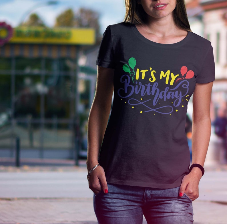 May include: Black t-shirt with the text "IT'S MY Birthday" in a colorful design featuring balloons. The text is in yellow, purple, and blue, with green and red balloons. The shirt is worn by a person in blue jeans.