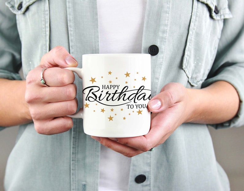 May include: White ceramic mug with the text "Happy Birthday to You" in black script and gold stars. The mug is held by a person wearing a light green denim jacket.