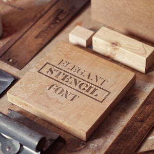 Puede incluir: Un bloque de madera con las palabras "Elegant Stencil Font" grabadas en &eacute;l. El bloque est&aacute; rodeado de otros objetos de madera y herramientas sobre una superficie de madera.