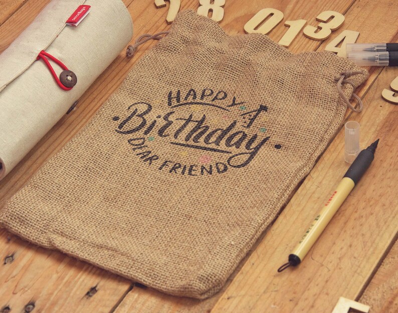 May include: A brown burlap drawstring bag with the words "Happy Birthday Dear Friend" in black script. A rolled-up beige fabric case with a red button closure is to the left. A pen and wooden numbers are also visible.