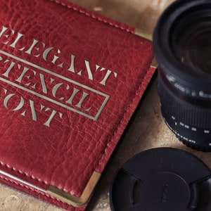 Puede incluir: Una libreta de cuero roja con detalles dorados y el texto "Elegant Stencil Font" en relieve en la portada. La libreta est&aacute; junto a un objetivo de c&aacute;mara negro y una tapa de objetivo negra.
