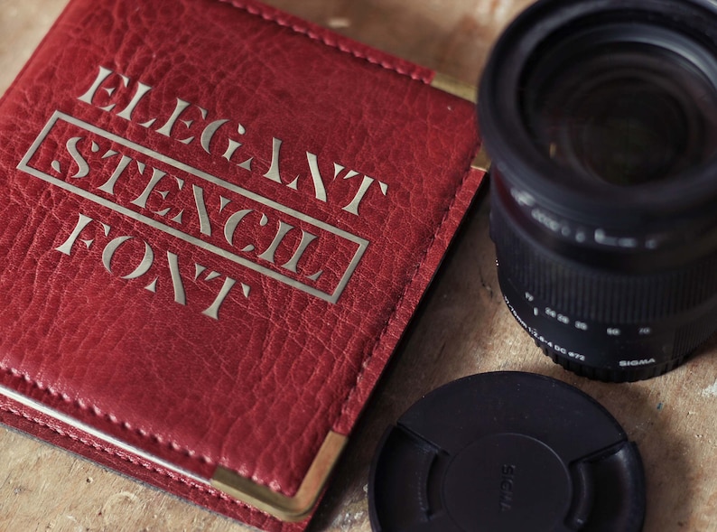 Puede incluir: Una libreta de cuero roja con detalles dorados y el texto "Elegant Stencil Font" en relieve en la portada. La libreta est&aacute; junto a un objetivo de c&aacute;mara negro y una tapa de objetivo negra.