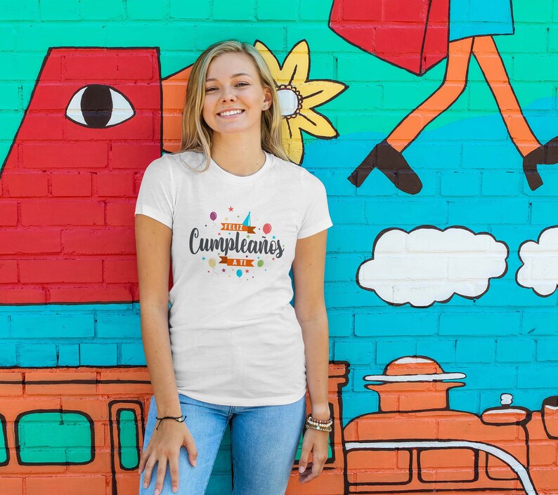 May include: A white t-shirt with the text "Feliz Cumplea&ntilde;os a ti" in black script, decorated with colorful confetti and a party hat. The shirt is worn by a person in blue jeans, standing in front of a colorful mural.