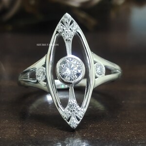 May include: A silver ring with a large, round, clear gemstone in the center. The ring has a delicate, openwork design with smaller gemstones on the sides. The ring is set on a dark background.