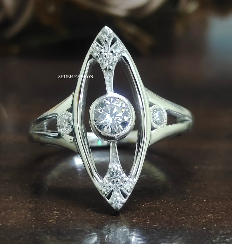 May include: A silver ring with a large, round, clear gemstone in the center. The ring has a delicate, openwork design with smaller gemstones on the sides. The ring is set on a dark background.