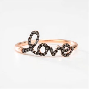 以下が含まれることがあります： ローズゴールドのリングに、黒いダイヤモンドで「love」の文字が書かれています。