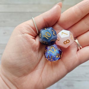 Könnte beinhalten: Drei polyedrische Würfel in einer Hand. Einer ist marineblau mit goldenen Zahlen, ein anderer ist hellrosa mit goldenen Zahlen, und der dritte ist tiefblau mit schillerndem Glitzer und goldenen Zahlen. Die Würfel sind für Tabletop-Spiele.
